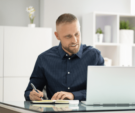 Man takes notes with pen and paper while looking at a laptop.